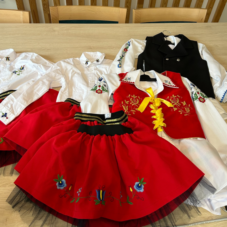 Na zdjęciu na jasnym, drewnianym stole leży kilka dziecięcych strojów ludowych. Składają się z białych koszul z długim rękawem, ozdobionych kolorowym haftem roślinnym na rękawach i przy kołnierzykach. Do koszul dołączone są czerwone spódniczki z haftowanymi wzorami kwiatowymi w dolnej części. Jeden strój zawiera czerwony gorset z żółtą tasiemką i złotym haftem. Obok znajduje się biały strój z czarnym kamizelkowym żakietem. W tle widać kilka drewnianych krzeseł ustawionych przy stole.