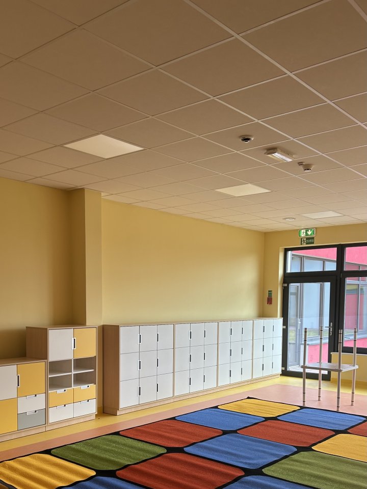 Sala przedszkolna urządzona w ciepłych barwach – dominują kolory żółty i pomarańczowy. W pomieszczeniu znajdują się niskie regały z zabawkami oraz małe stoliki i krzesełka dla dzieci w dopasowanych kolorach. Na podłodze leży duży, kolorowy dywan.