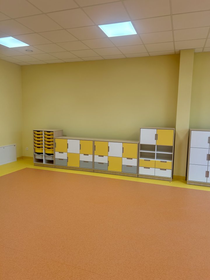 Sala przedszkolna urządzona w ciepłych kolorach – dominują odcienie żółtego i pomarańczowego. Na jednej ze ścian ustawiono niskie regały na zabawki.